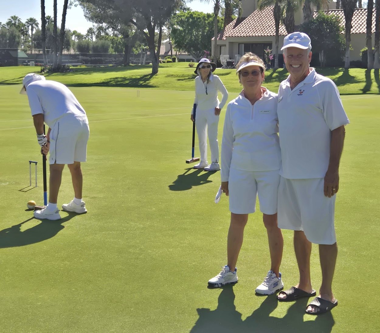 Two smiling members in whites on the lawn while a game plays on behind them
